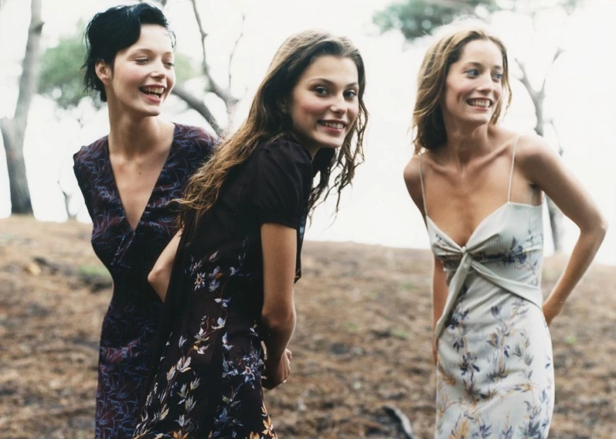 Three women in floral dresses stand closely together in a grassy outdoor setting with trees in the background, all smiling and relaxed.