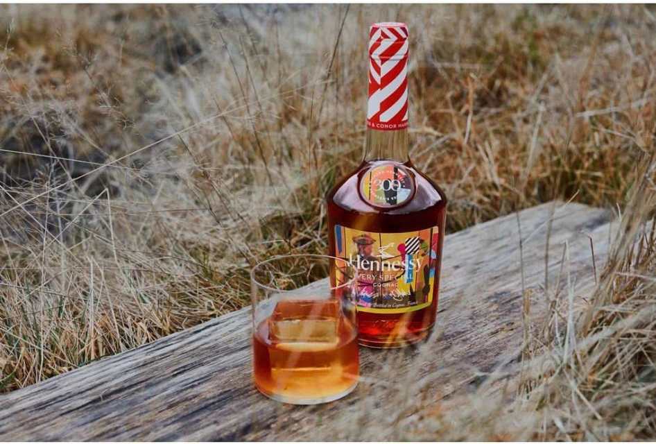 A liquor bottle and a glass with an ice cube sit on a wooden surface surrounded by tall, dry grass in an outdoor setting.