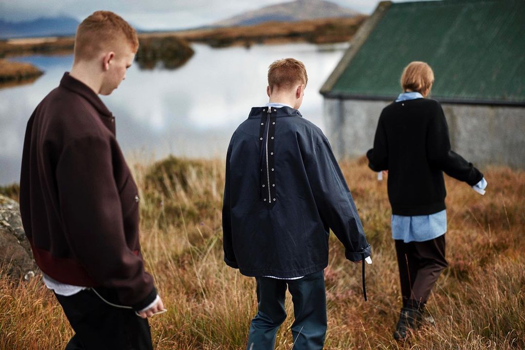 Three people walk through tall grass in a rural landscape with a small building and distant mountains under an overcast sky.