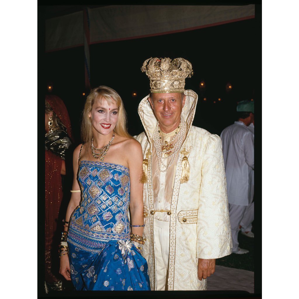 Blonde woman in a blue dress and a man in a gold crown and white robe pose together at an indoor event with dark lighting.