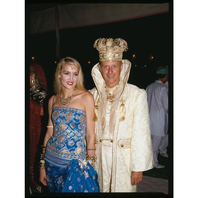 Blonde woman in a blue dress and a man in a gold crown and white robe pose together at an indoor event with dark lighting.