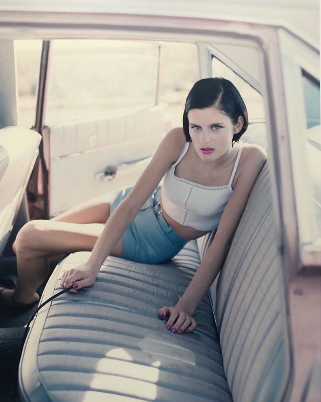 A woman in a white top and denim shorts lounges in the back seat of a vintage car with the door open in a sunlit outdoor setting.