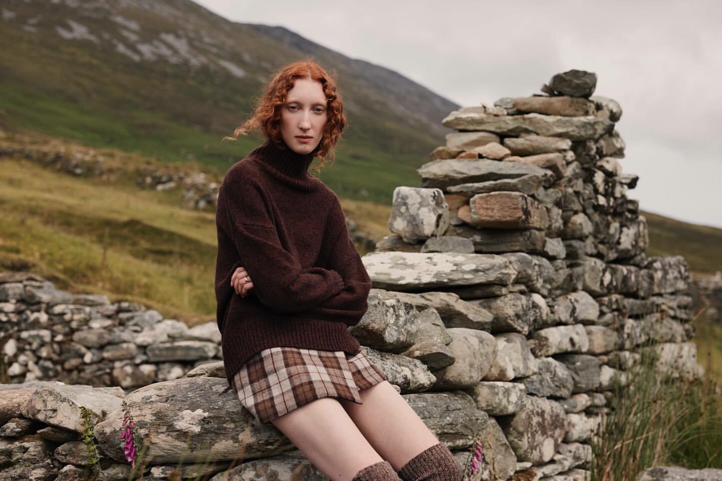 A red-haired young woman in a brown turtleneck sweater and plaid skirt poses sitting on a stone wall in a rural landscape.