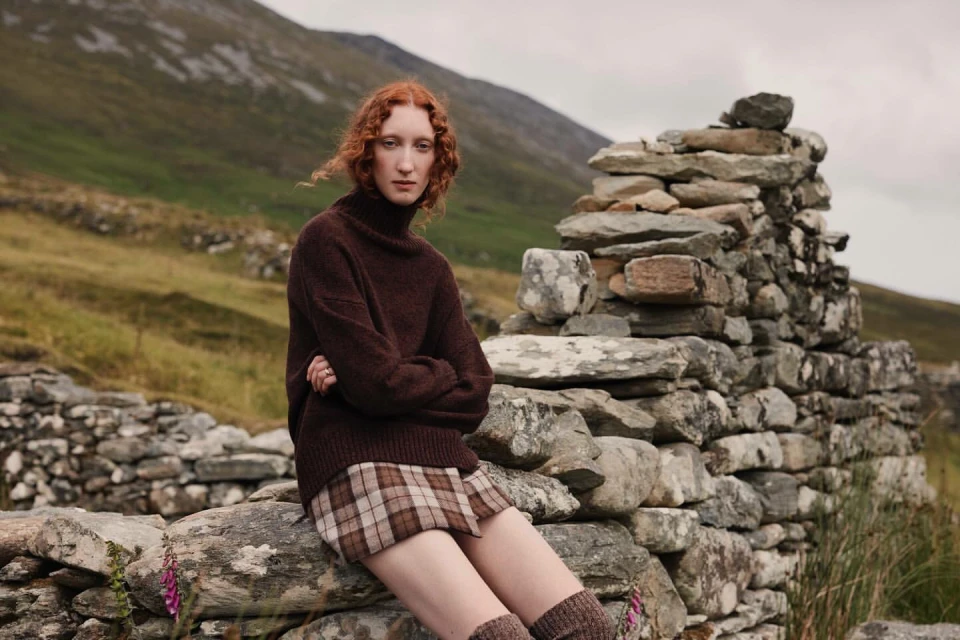 A red-haired young woman in a brown turtleneck sweater and plaid skirt poses sitting on a stone wall in a rural landscape.