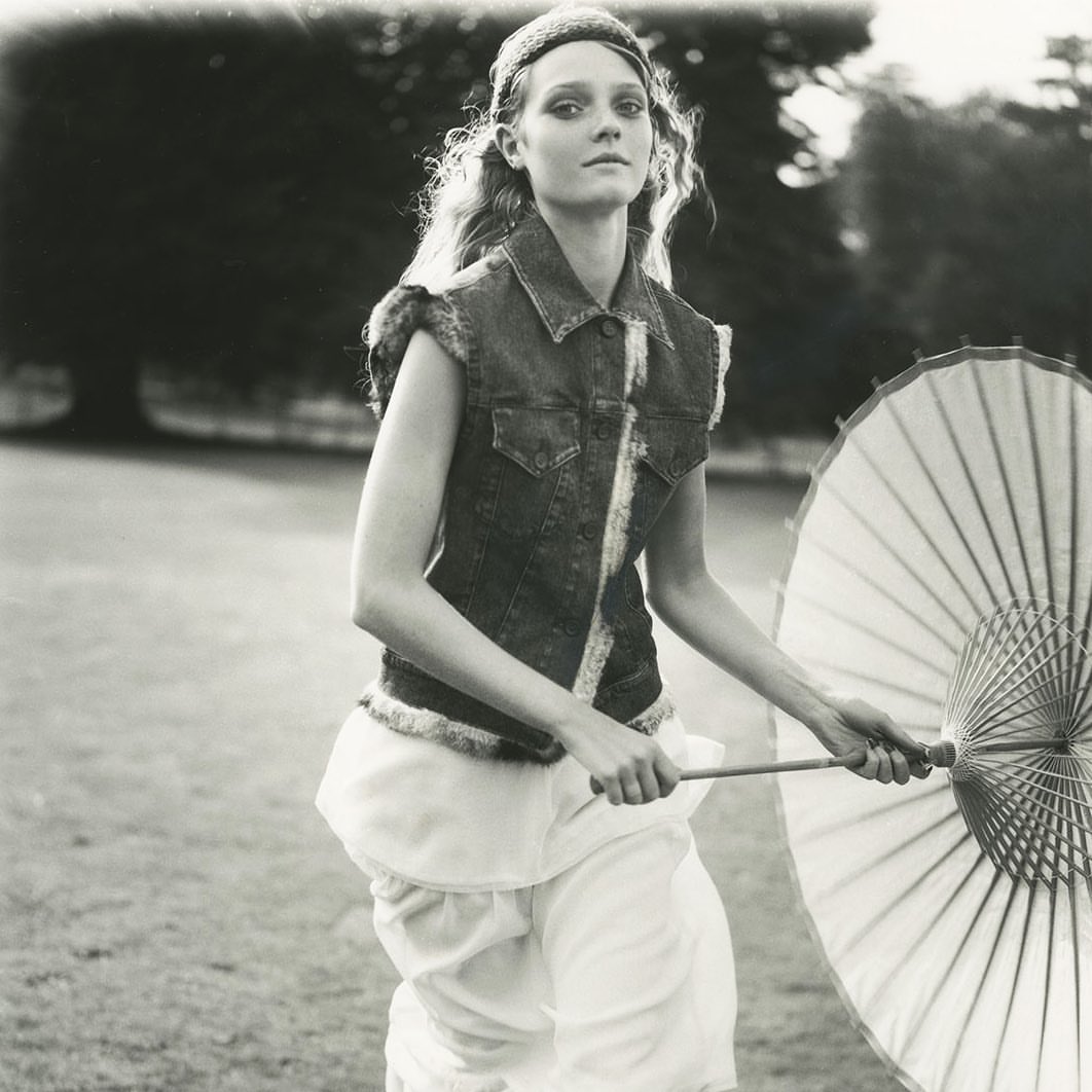 Black and white shot of a model in a denim vest and light skirt, holding an umbrella, outdoors in an open field on a sunny day.