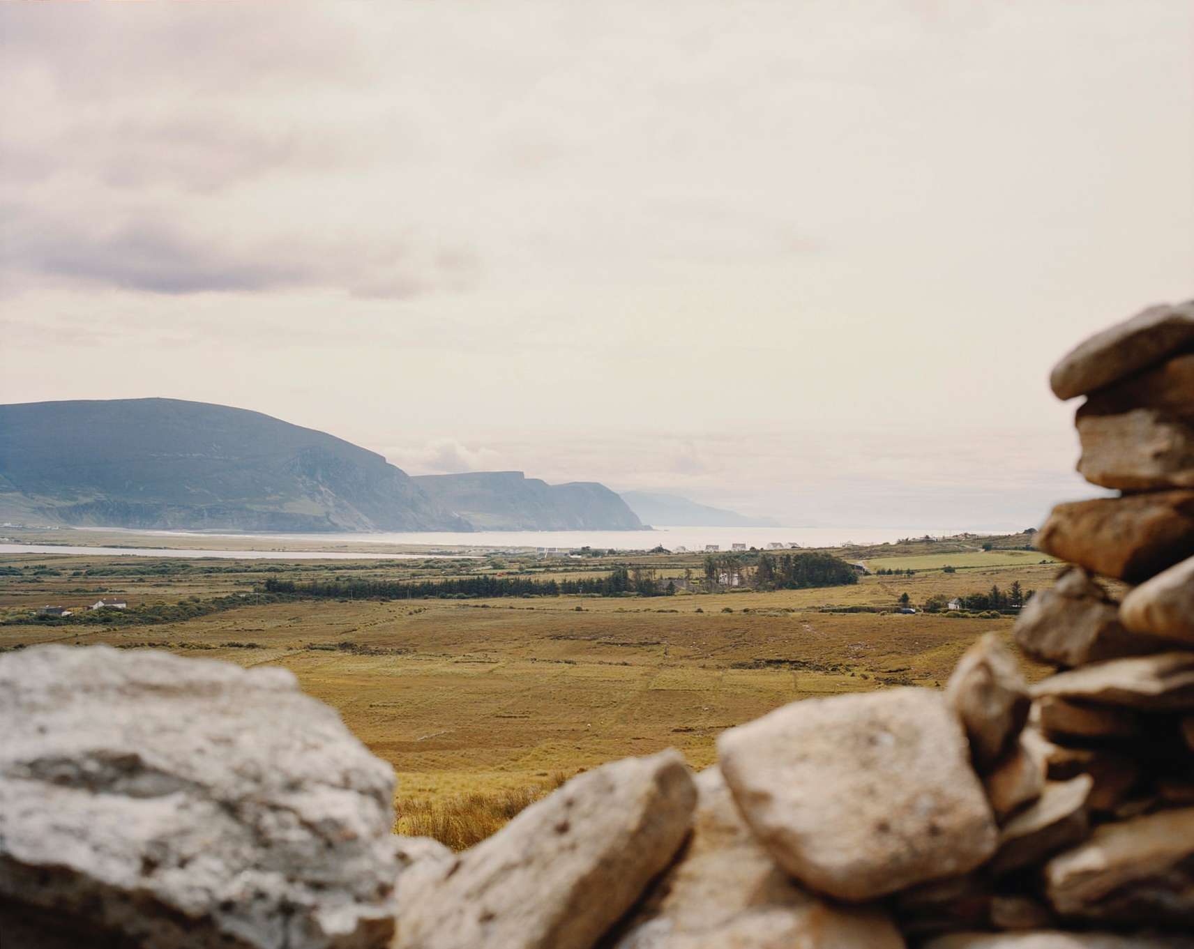 Scenic view of a coastal landscape with mountains and a body of water, framed by a stone wall in the foreground under an overcast sky.