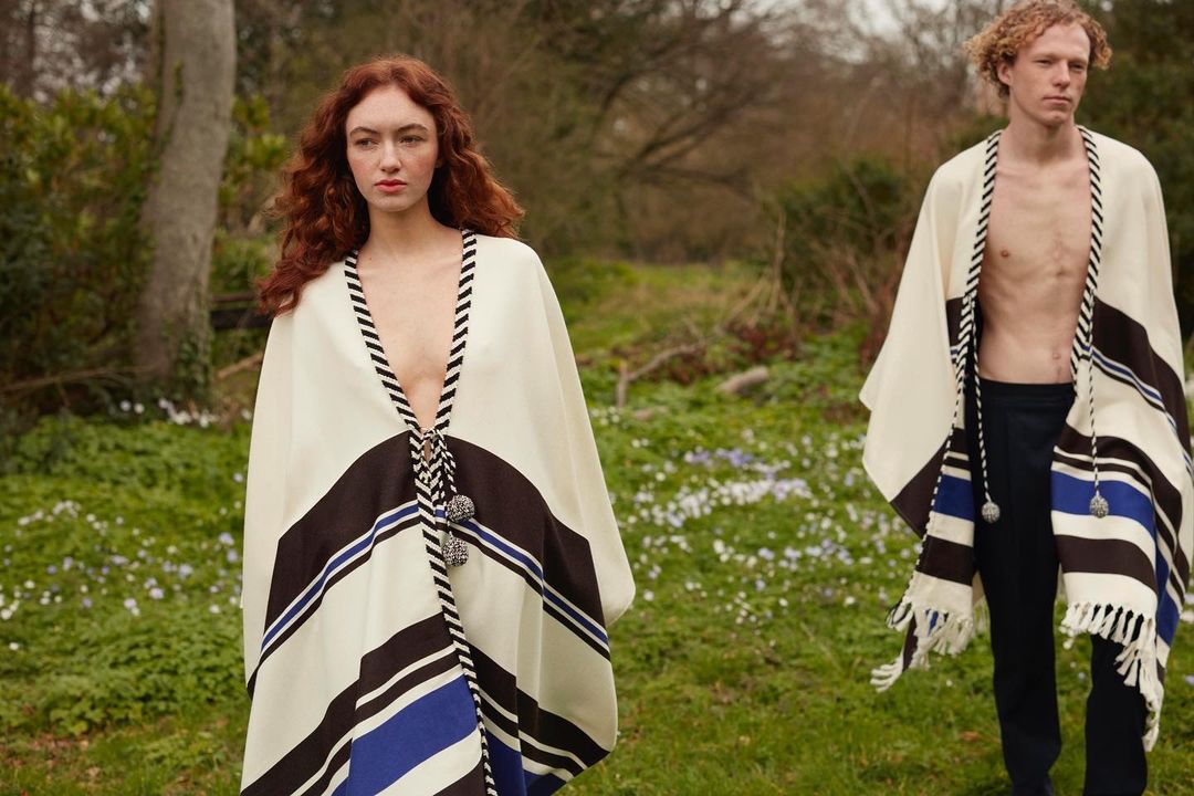 Two models pose outdoors in matching white and blue striped ponchos. One is a female with red hair; the other is a shirtless male.