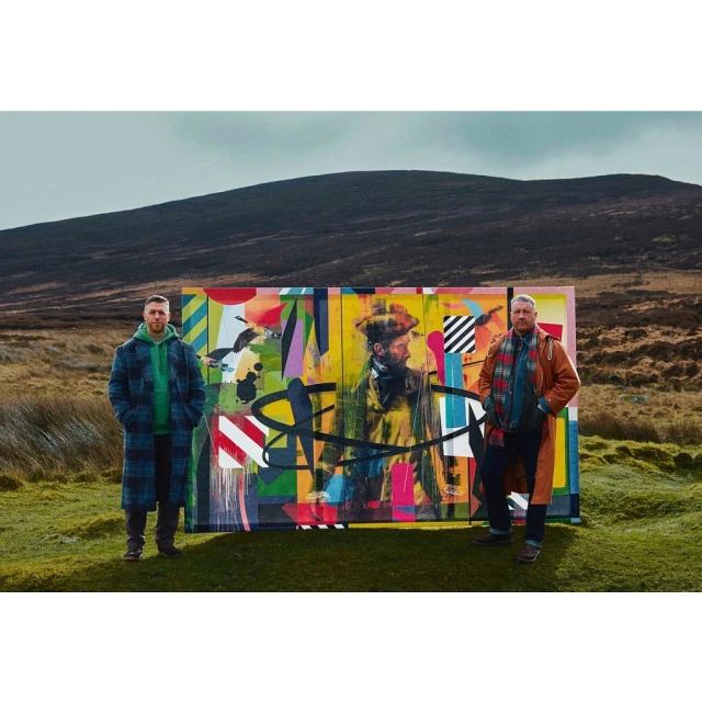 Two men posing with a colorful outdoor art installation in a grassy, hilly landscape under a cloudy sky, showcasing fashion and art.