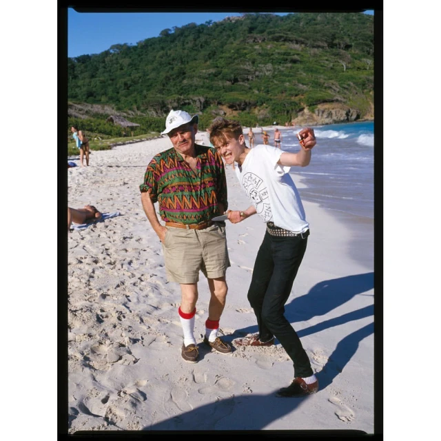 Two men pose on a sunny beach with white sand and turquoise water, backed by a green, hilly landscape. One man is pointing and smiling.