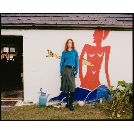 Red-haired model in blue polo and pleated skirt standing before a whitewashed wall with a large painted figure.