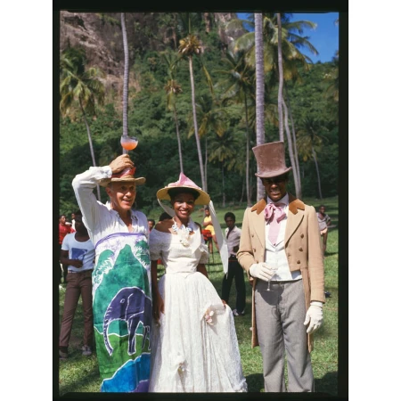 Three people in vintage clothing stand on a green lawn, with palm trees and mountains in the background, creating a festive outdoor scene.