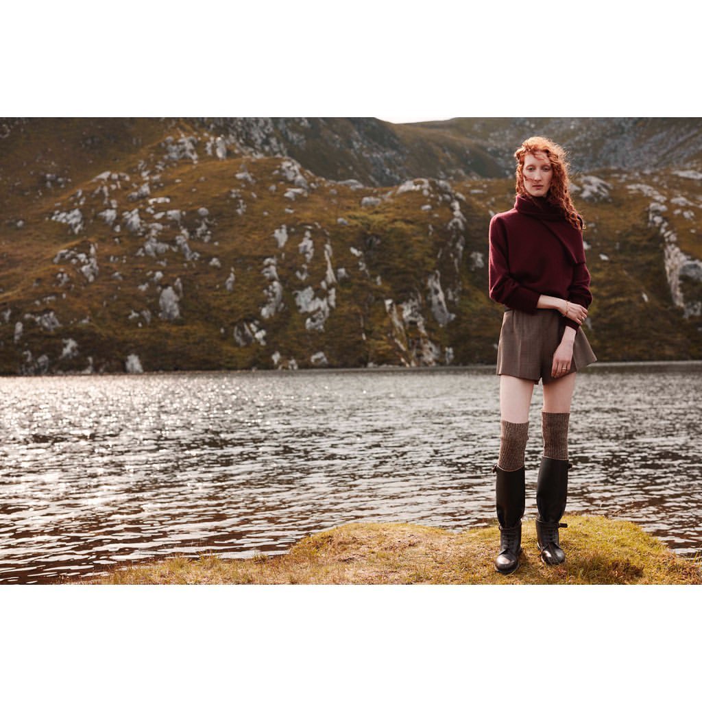 A woman with red hair poses in a burgundy turtleneck sweater, brown shorts, and black boots, standing in front of a lake and mountains.