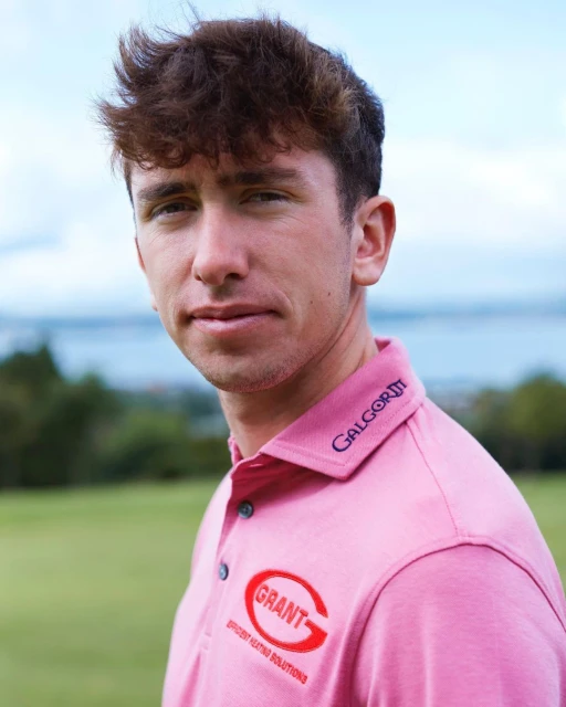 Close-up portrait of a smiling adult male with brown hair wearing a pink polo shirt in a green outdoor setting with a water view.