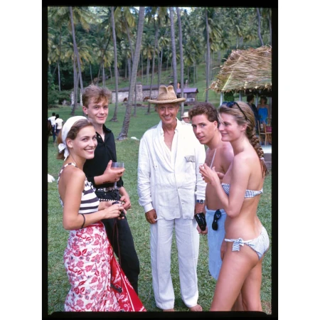 Group of people standing in a tropical setting with palm trees, a thatched roof structure, and lush greenery in the background.