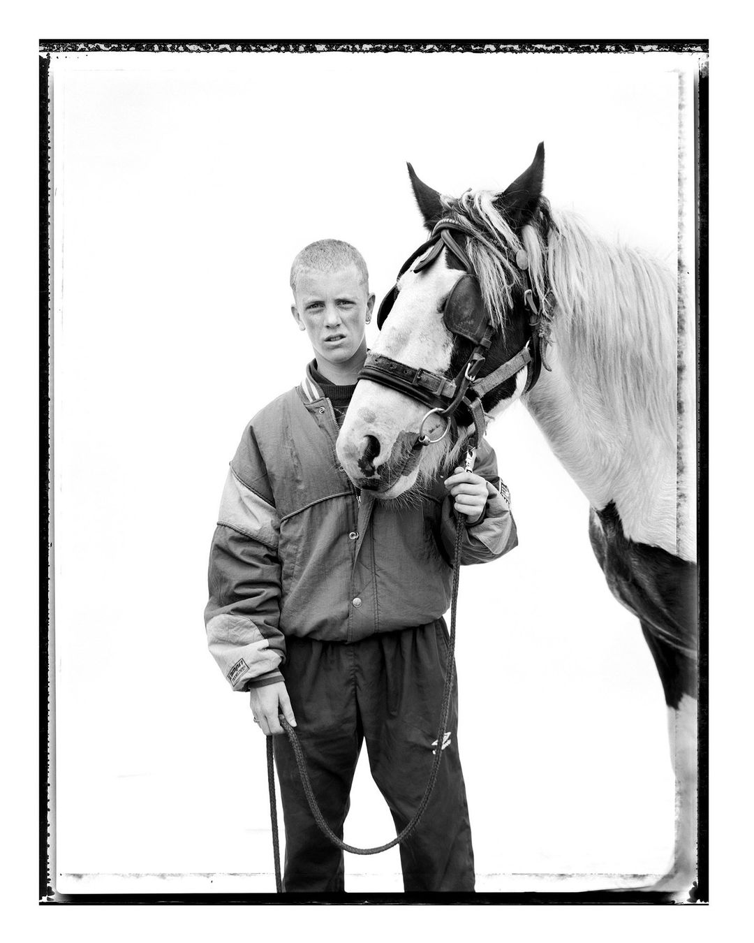 A black and white portrait showcases a young man in a jacket standing next to a horse with a bridle, set against a stark white backdrop.