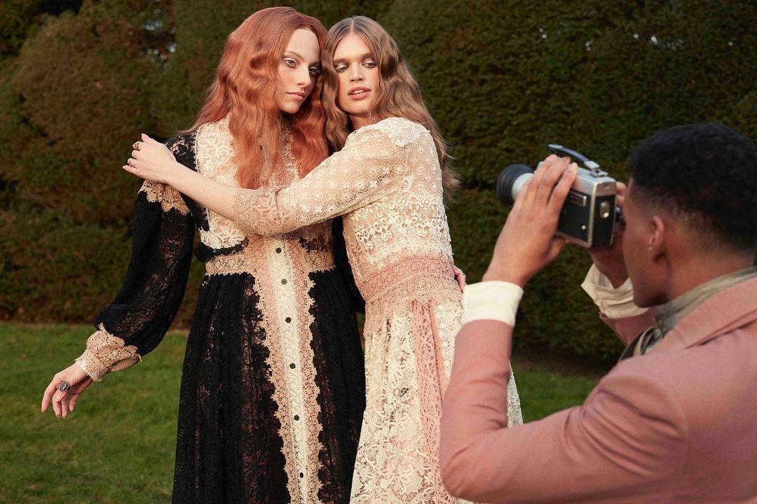 Two women in lace dresses pose while a photographer captures them with a vintage camera, set against a backdrop of greenery.