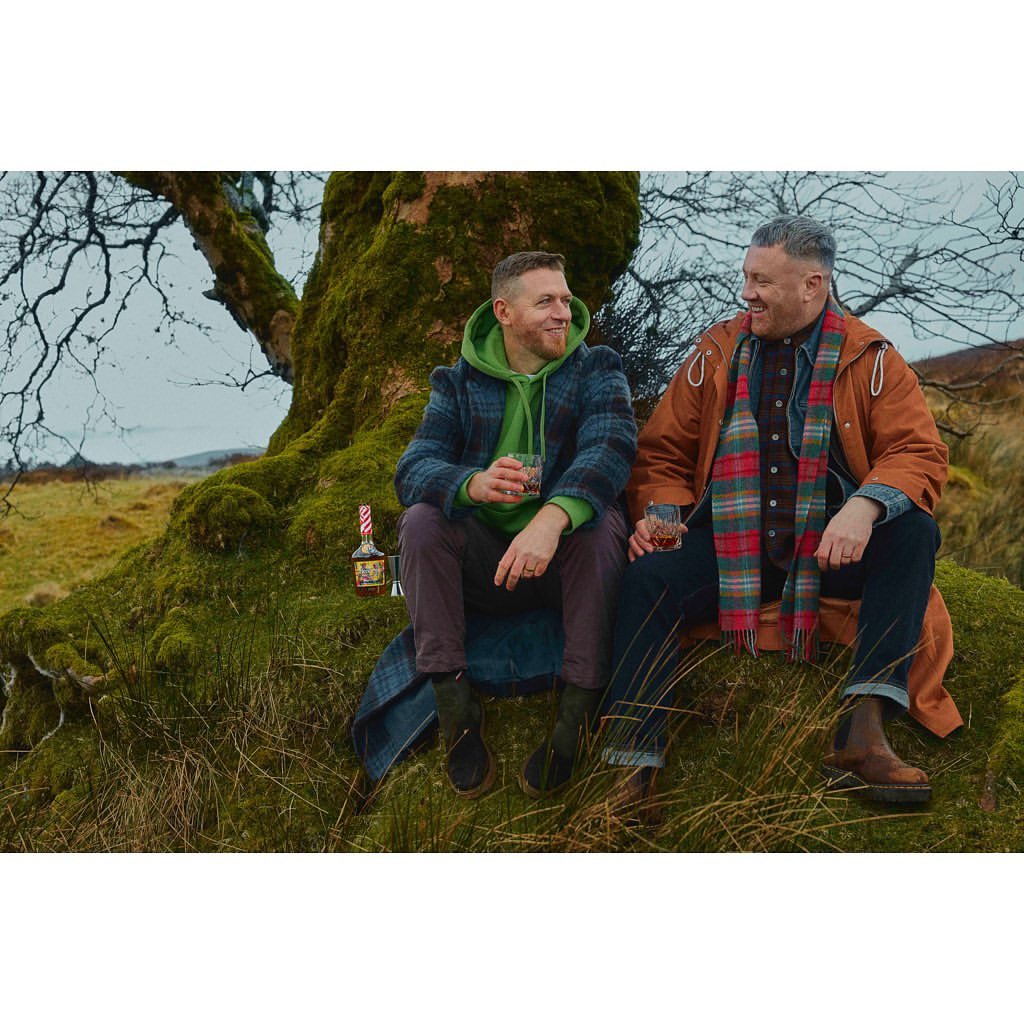 Two adult men sit outside with a bottle and glasses. They are sitting on a mossy rock near a tree in a grassy field.