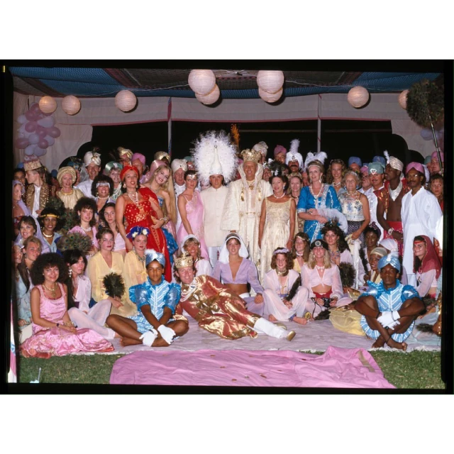 Large group of people dressed in costumes with headdresses gather outdoors under tents with paper lanterns for a festive occasion.