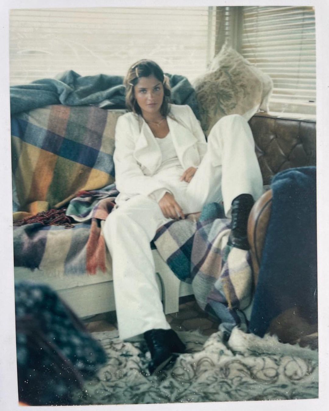 A woman in a white suit poses on a couch with plaid blankets near a window, looking at the camera in a medium shot.