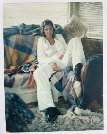 A woman in a white suit poses on a couch with plaid blankets near a window, looking at the camera in a medium shot.