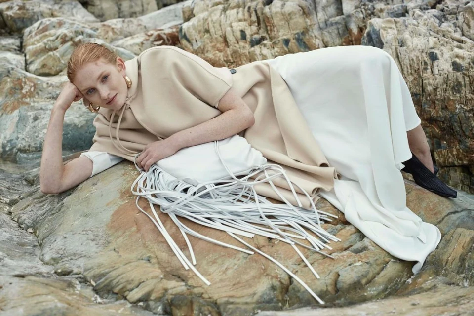 Red-haired fashion model lies on coastal rocks in a beige top, long white skirt, gold hoop earrings and a fringe bag, outdoors.