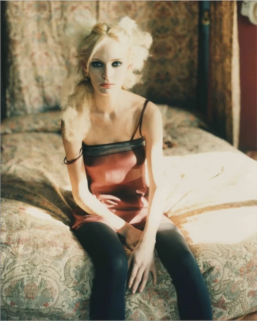 Portrait of a blonde woman with an ethereal aesthetic sitting on a bed with patterned sheets and a thin-strapped dress in soft light.