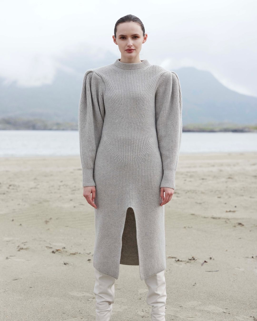 A woman poses on a sandy beach wearing a grey sweater dress and white boots, with mountains and water in the background on an overcast day.