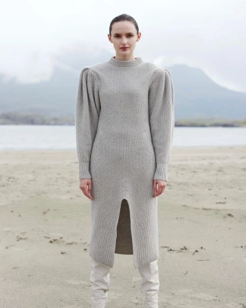 A woman poses on a sandy beach wearing a grey sweater dress and white boots, with mountains and water in the background on an overcast day.