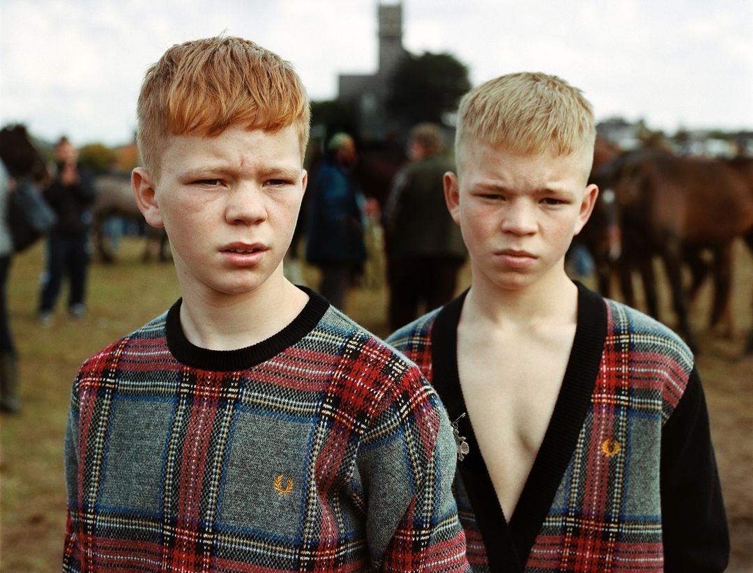 Portrait of two boys with red hair, wearing matching plaid sweaters, standing in an outdoor setting with horses and people.