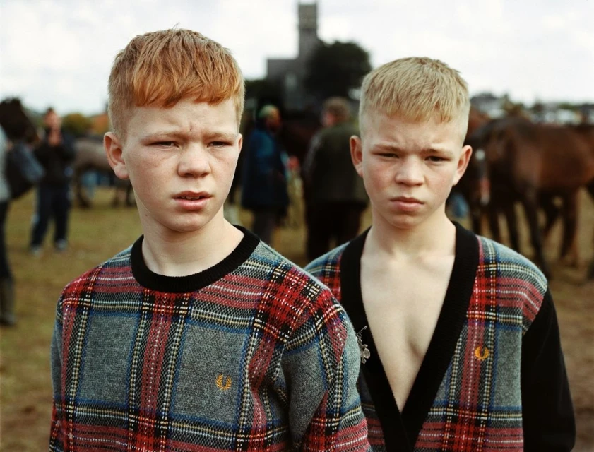 Portrait of two boys with red hair, wearing matching plaid sweaters, standing in an outdoor setting with horses and people.
