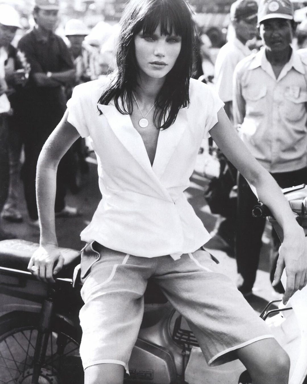 Monochrome fashion shot of a woman in a white top and shorts sitting on a motorbike surrounded by a crowd of people.