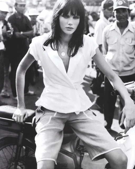 Monochrome fashion shot of a woman in a white top and shorts sitting on a motorbike surrounded by a crowd of people.