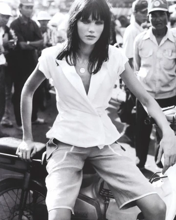 Monochrome fashion shot of a woman in a white top and shorts sitting on a motorbike surrounded by a crowd of people.
