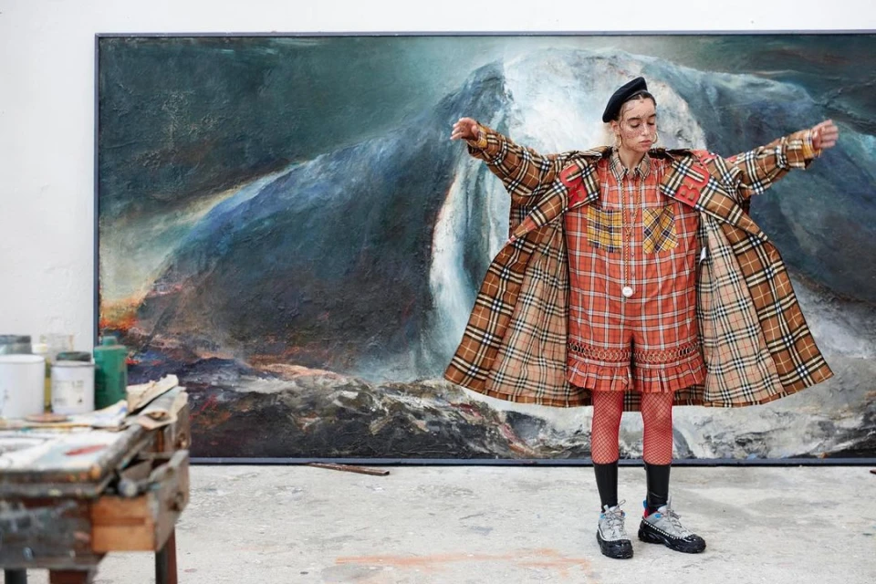 Woman in plaid outfit poses in front of large painting in an art studio, wearing beret and chunky sneakers, arms outstretched.