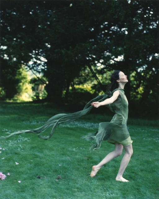 Woman in green dress runs barefoot through a grassy field with trees in the background, holding a scarf fluttering behind her.