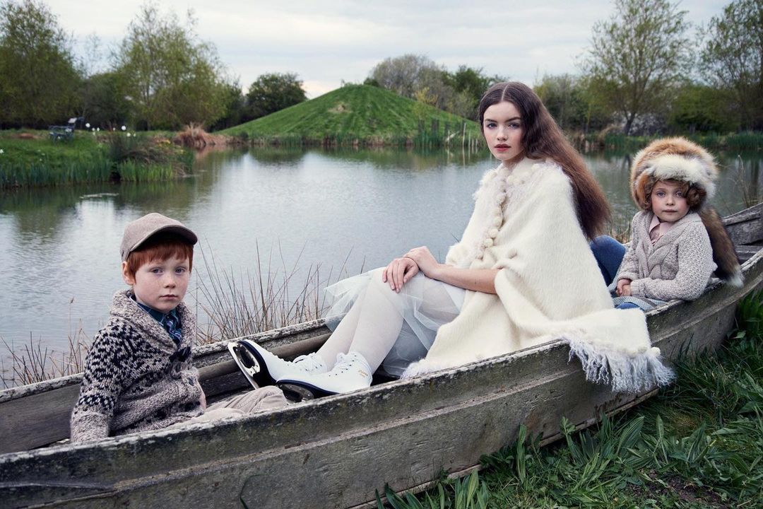 Two children and an adult female are sitting in a small boat on a lake with a grassy hill in the background, wearing vintage clothing.