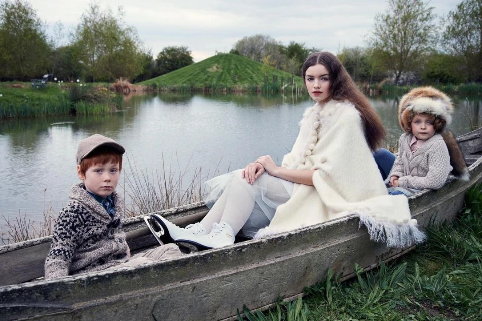Two children and an adult female are sitting in a small boat on a lake with a grassy hill in the background, wearing vintage clothing.