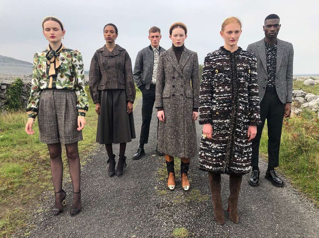 Five models pose on a path outdoors, showcasing a collection of tweed coats, jackets, and skirts in neutral tones, blending fashion with nature.