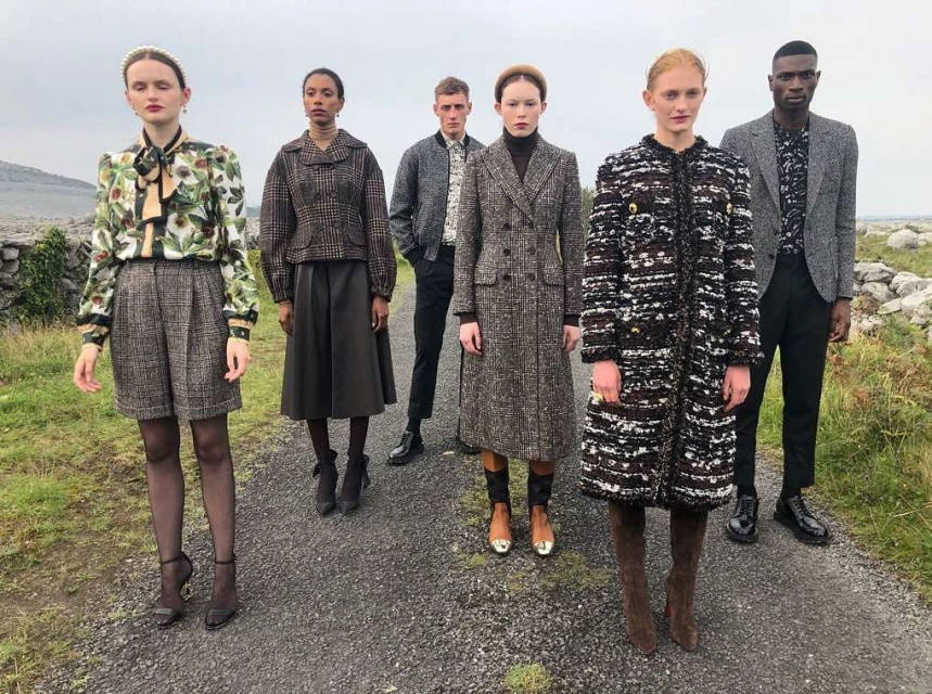 Five models pose on a path outdoors, showcasing a collection of tweed coats, jackets, and skirts in neutral tones, blending fashion with nature.