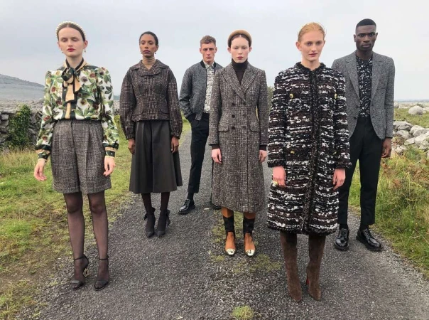 Five models pose on a path outdoors, showcasing a collection of tweed coats, jackets, and skirts in neutral tones, blending fashion with nature.