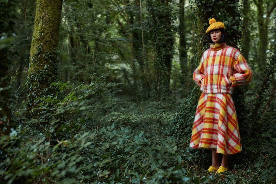 Woman in plaid sweater and skirt posing in forest. She wears a yellow beret and shoes among the green trees in the natural setting.