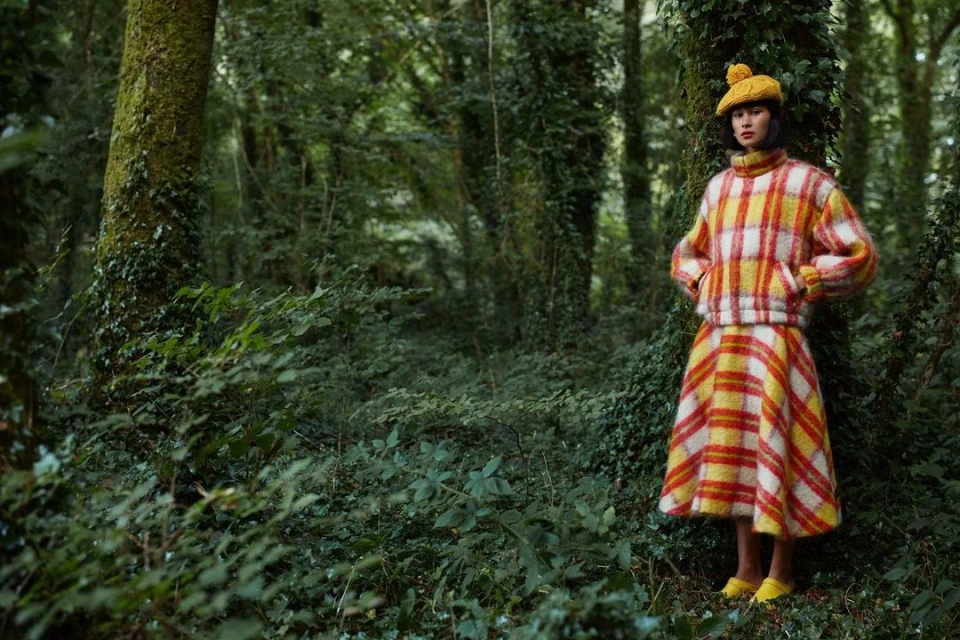 Woman in plaid sweater and skirt posing in forest.  She wears a yellow beret and shoes among the green trees in the natural setting.