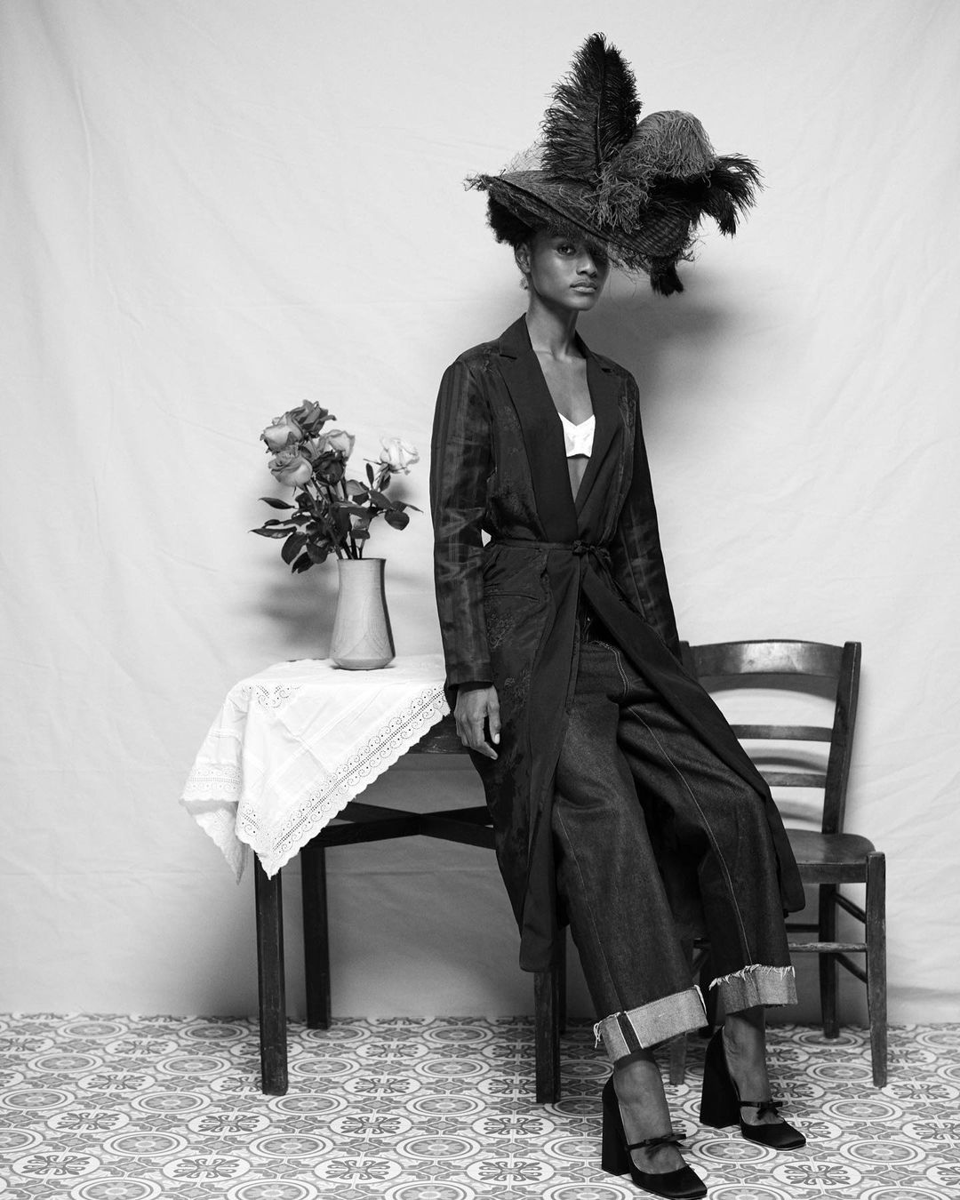 Elegant fashion portrait of a Black woman in a large feathered hat, long coat, and platform shoes, seated in a photo studio.