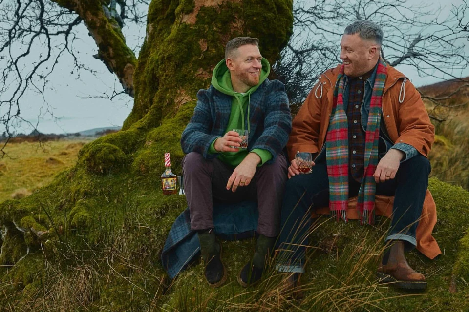 Two adult males enjoy whiskey outdoors. They are sitting on a mossy tree in a green field beneath a cloudy sky, showcasing outdoor lifestyle.