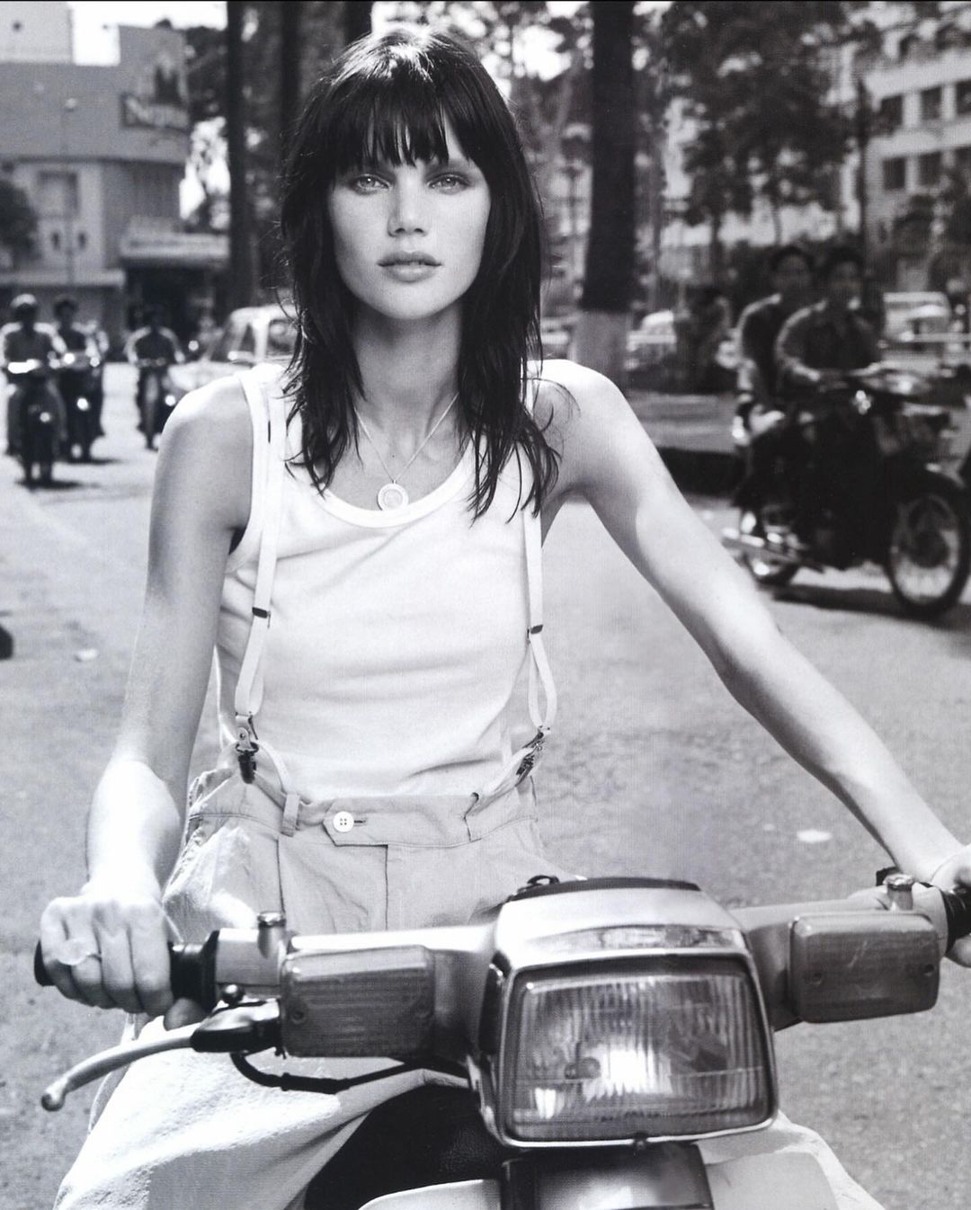 Black-and-white portrait of a brunette woman riding a motor scooter on a city street, showcasing fashion and urban life.