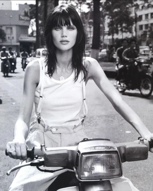 Black-and-white portrait of a brunette woman riding a motor scooter on a city street, showcasing fashion and urban life.