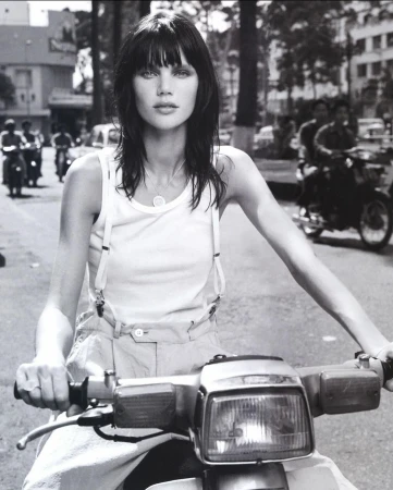 Black-and-white portrait of a brunette woman riding a motor scooter on a city street, showcasing fashion and urban life.