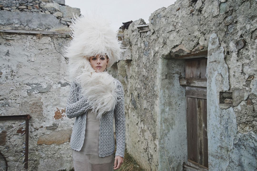 Woman in oversized feathered hat and grey knit cardigan stands in front of weathered stone structure, creating a surreal fashion image.