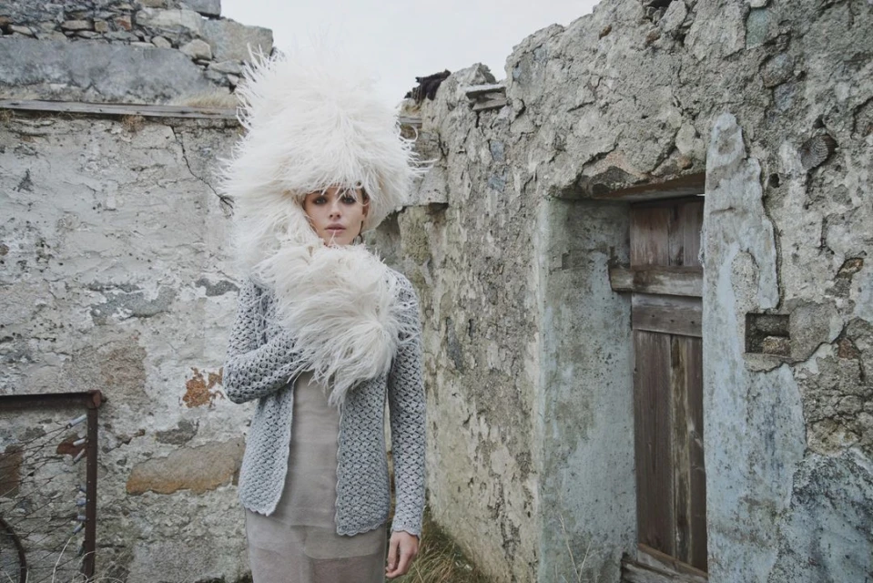 Woman in oversized feathered hat and grey knit cardigan stands in front of weathered stone structure, creating a surreal fashion image.