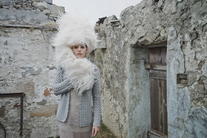 Woman in oversized feathered hat and grey knit cardigan stands in front of weathered stone structure, creating a surreal fashion image.
